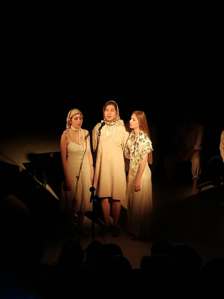 Półmrok, ciepłe żółte światło pada na stojące w półokręgu śpiewające trzy młode kobiety w jasnych długich sukniach stylizowanych na ludowe. Dwie z kobiet noszą na głowie chusty, chusta trzeciej jest zarzucona na ramiona.