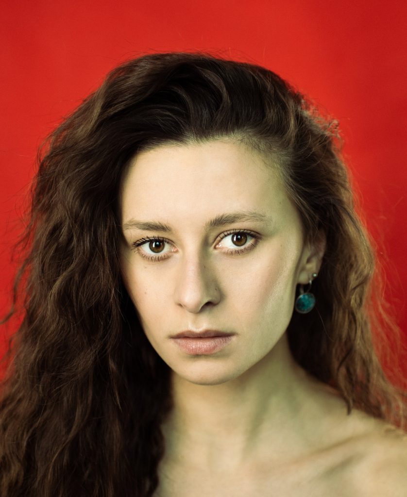 na czerwonym tle ujęcie portretowe młodej kobiety z brązowymi oczami i długimi ciemnymi włosami.