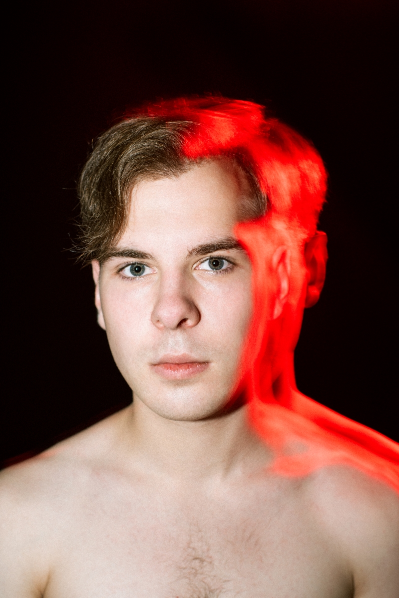 Portret młodego niebieskookiego blondyna. Światło padające na tył głowy i nagie ramiona mężczyzny wyraźnie kontrastuje z czarnym tłem i tworzy intensywną czerwoną poświatę.