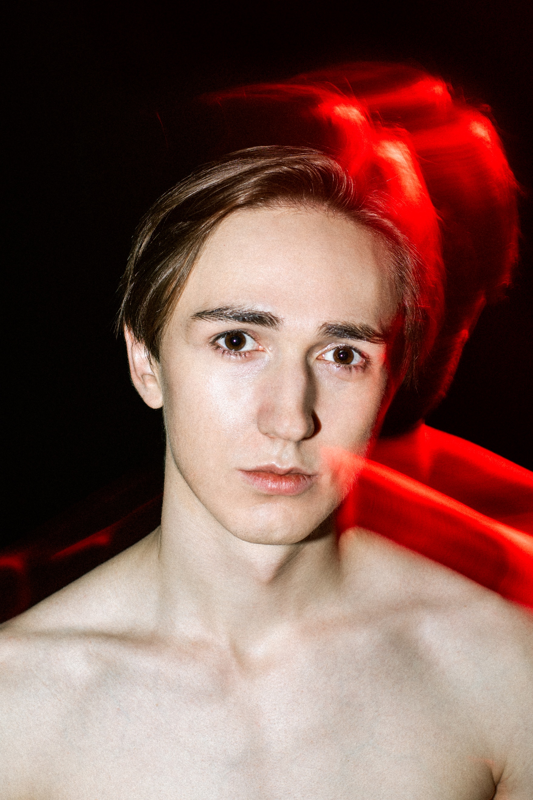 Portret młodego brązowookiego mężczyzny. Światło padające na tył głowy i nagie ramiona mężczyzny wyraźnie kontrastuje z czarnym tłem i tworzy intensywną czerwoną poświatę.