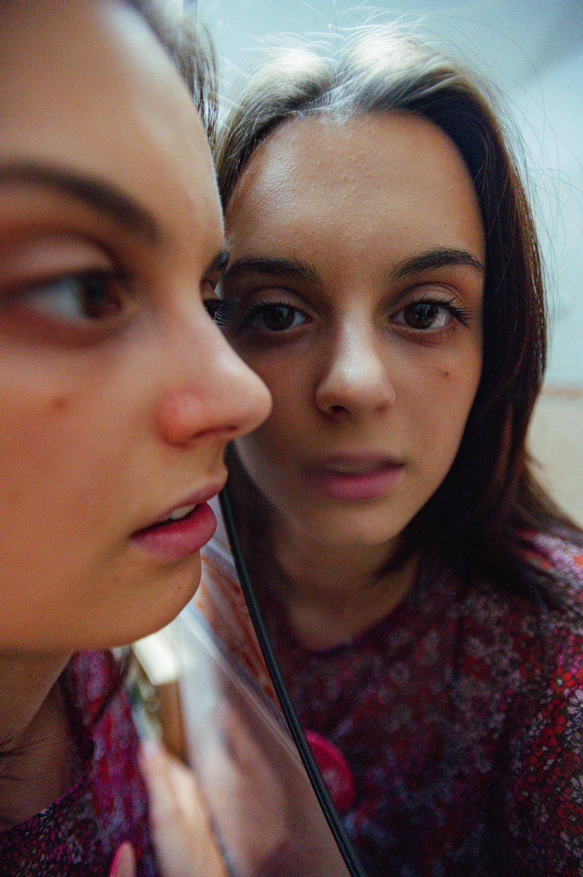 Młoda brunetka we wzorzystej różowoczerwonej bluzce przykłada bokiem swoją twarz do lustra, która się w nim odbija.