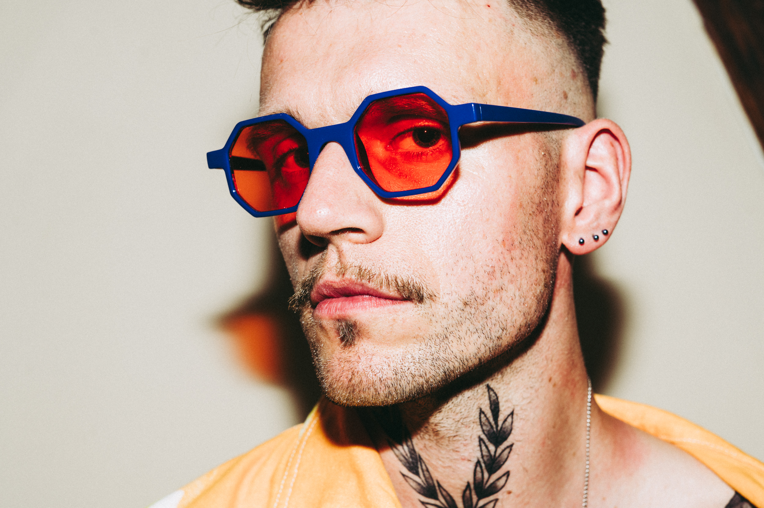Portret. Młody mężczyzna w okularach z czerwonopomarańczowymi szkłami i ciemnoniebieskimi oprawkami. W uchu ma trzy niewielkie, kuliste kolczyki, a na szyi tatuaż przedstawiający dwie splecione gałązki.