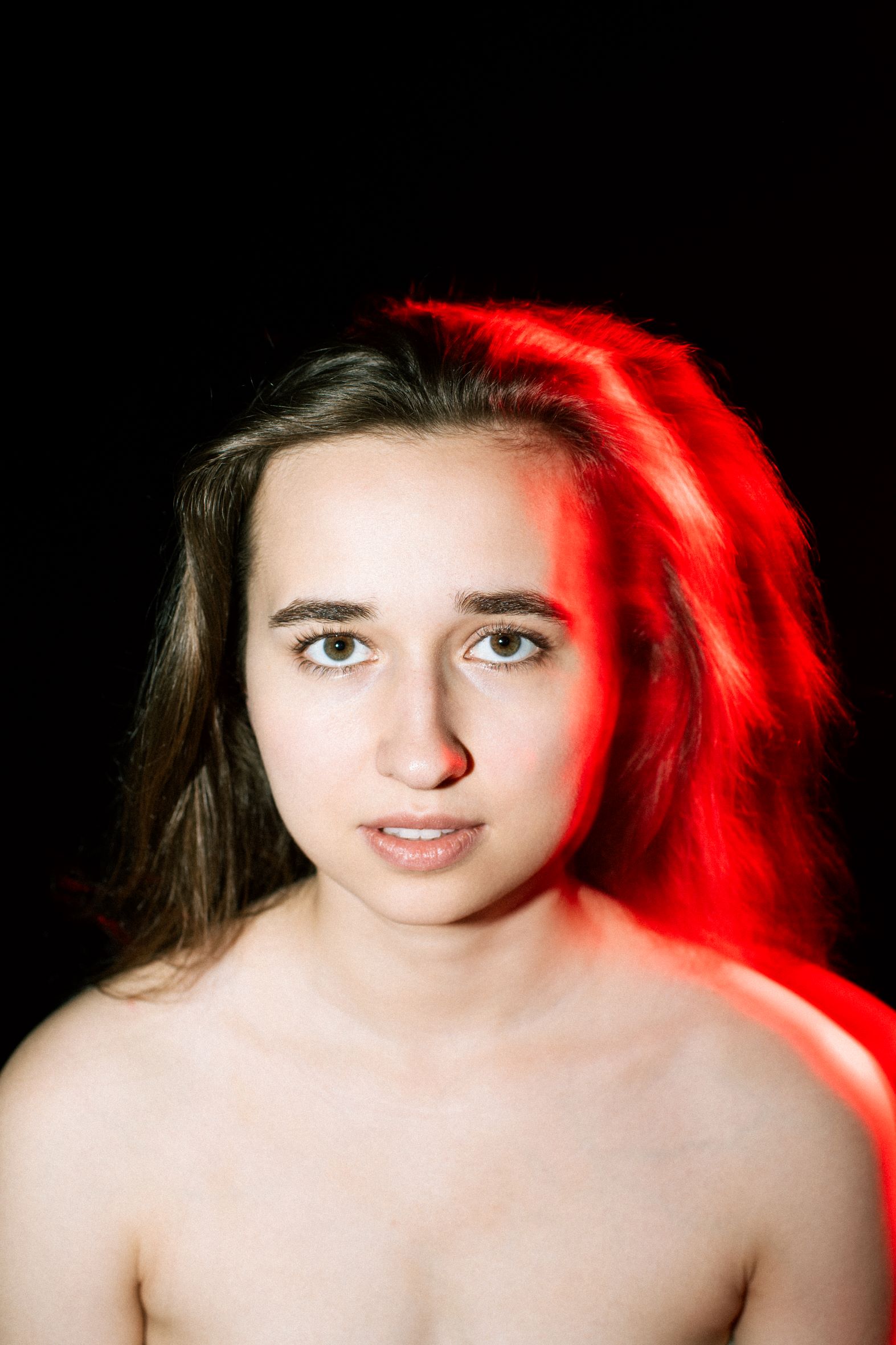 Portret młodej długowłosej blondynki. Światło padające z tyłu na głowę i nagie ramiona kobiety wyraźnie kontrastuje z czarnym tłem i tworzy intensywną czerwoną poświatę.