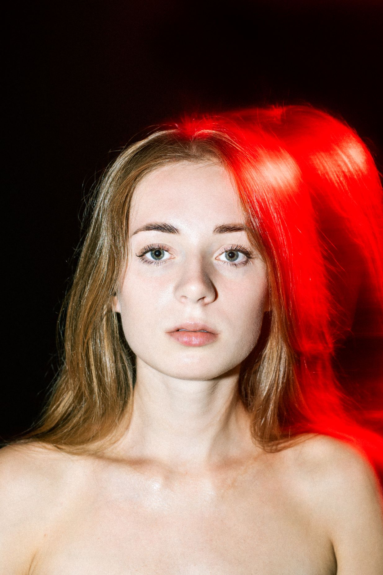 Portret młodej długowłosej blondynki z niebieskimi oczami. Światło padające z tyłu na głowę i nagie ramiona kobiety wyraźnie kontrastuje z czarnym tłem i tworzy intensywną czerwoną poświatę.
