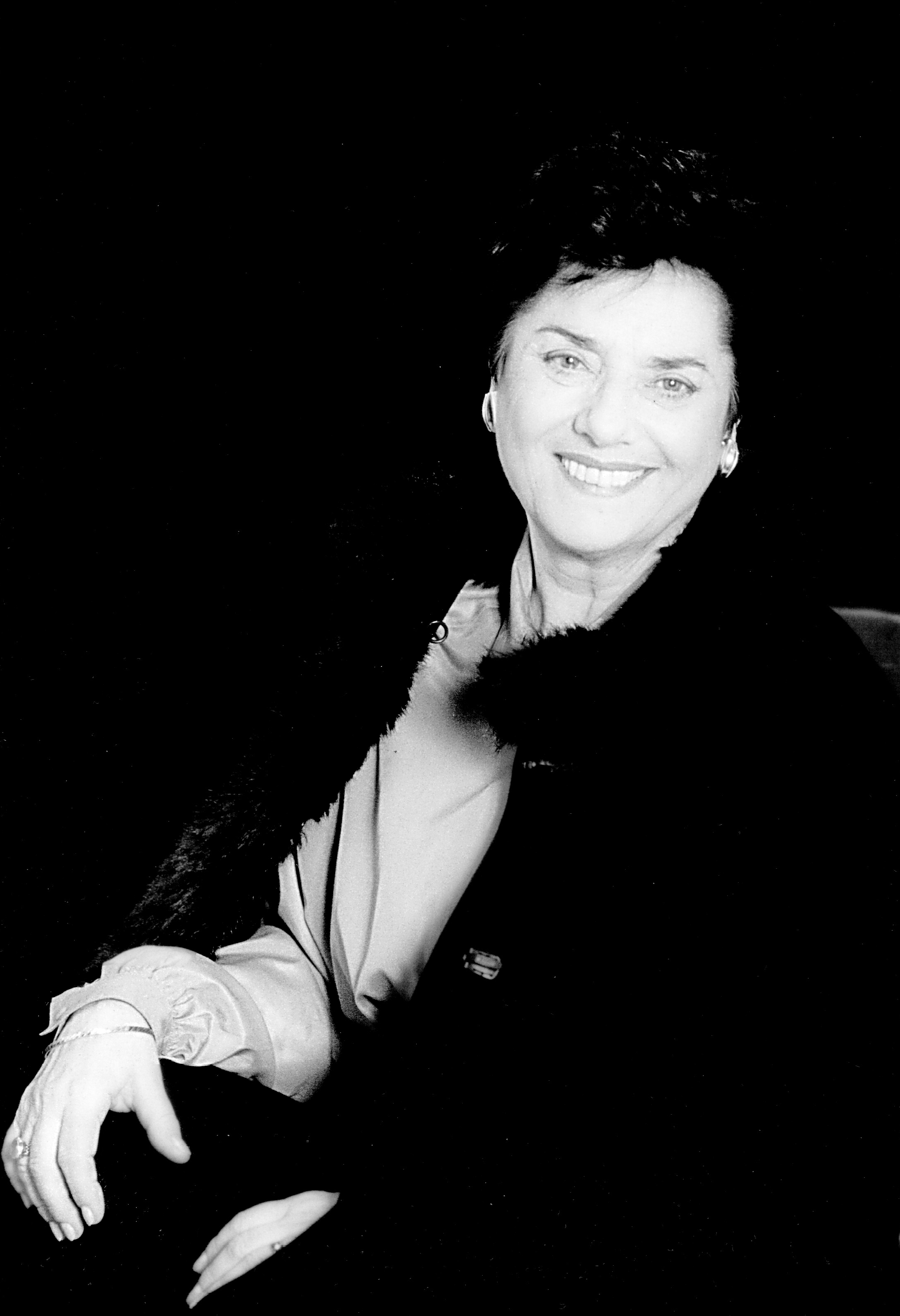 Czarnobiały portret kobiety w średnim wieku. Ma ciemne krótkie włosy, ubrana w jasną bluzkę koszulową i płaszcz z futrzanym kołnierzem. W uszach kolczyki, na rękach bransoletki i pierścionki.
