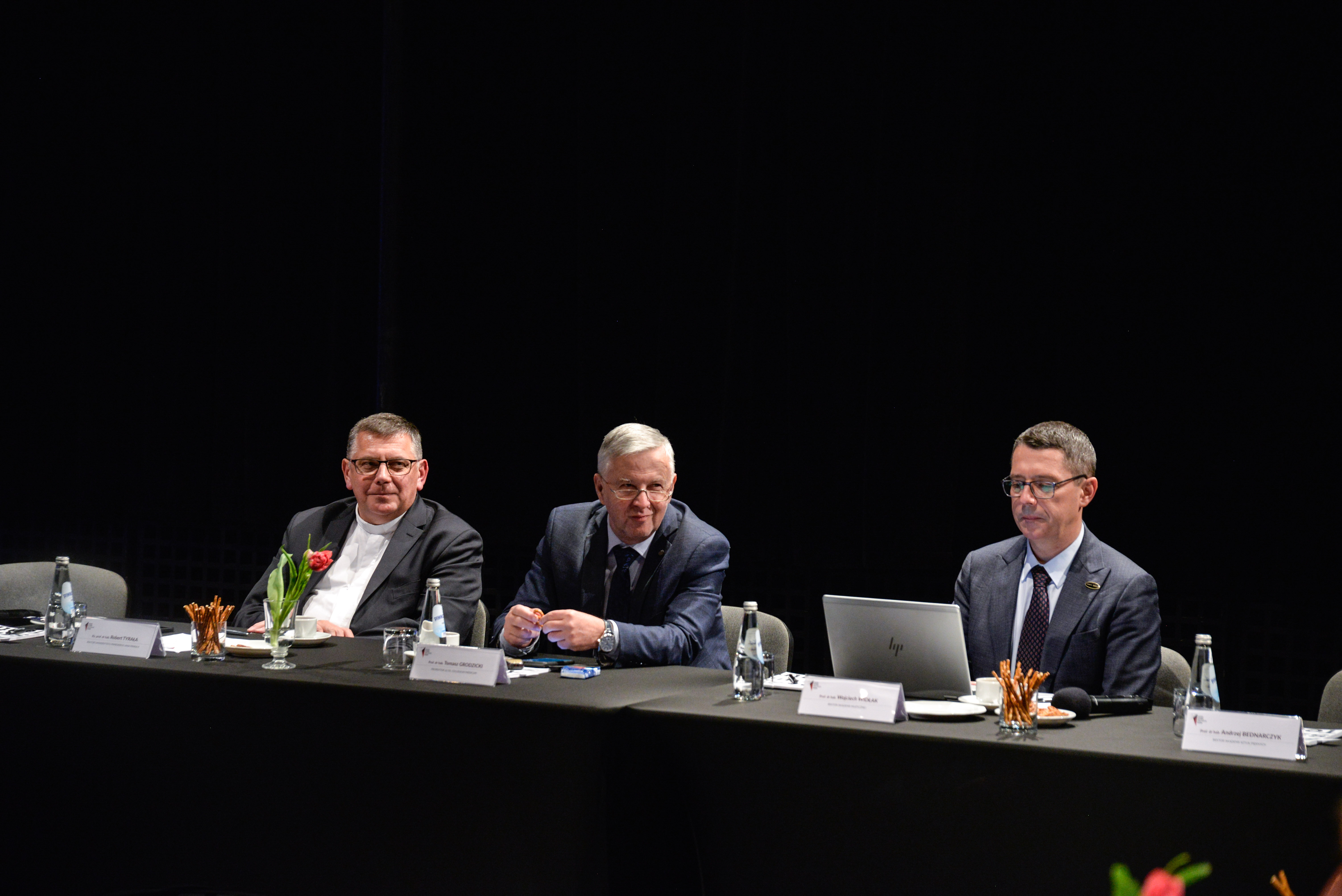 Obrady Kolegium Rektorów Szkól Wyższych Krakowa, przy nakrytym czarnym obrusem stołach siedzi trzech elegancko ubranych mężczyzn.