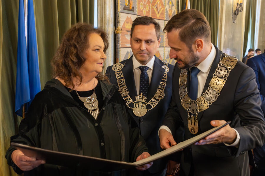 Kobieta i dwóch mężczyzn. Kobieta ubrana w czarną togę i złoty ozdobny naszyjnik, mężczyźni w eleganckich garniturach ze złotymi łańcuchami z herbem Krakowa na szyjach przeglądają wspólnie dokument w otwartej teczce.
