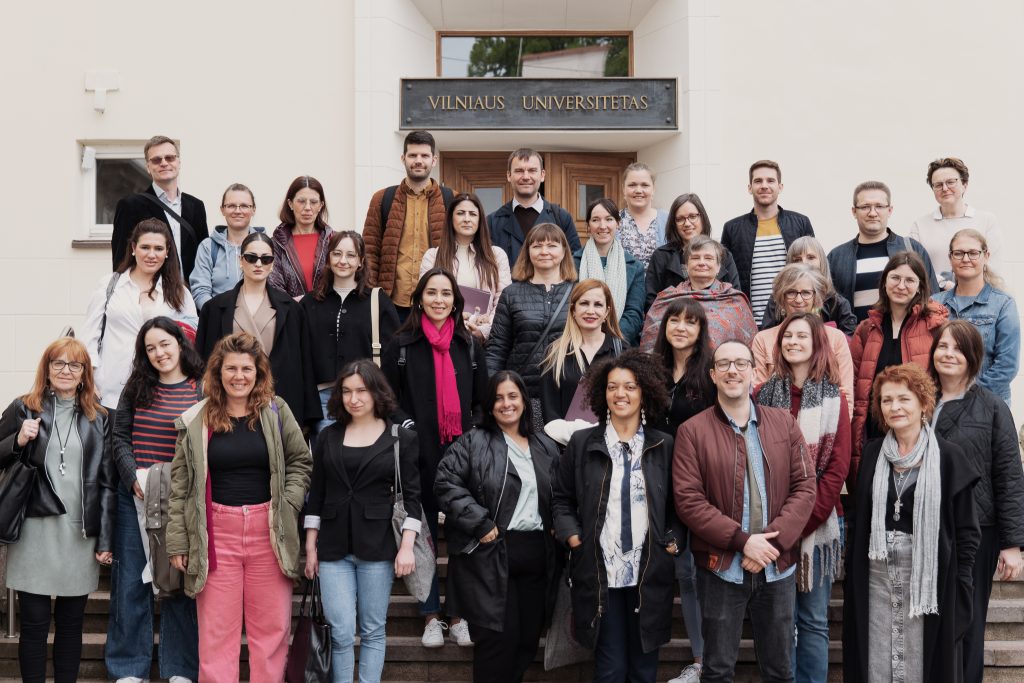 Na zewnątrz. Zróżnicowana grupa ponad 30 osób stojących przed budynkiem Vilnius University. Nad nimi nazwa tablica z nazwą uniwersytetu.