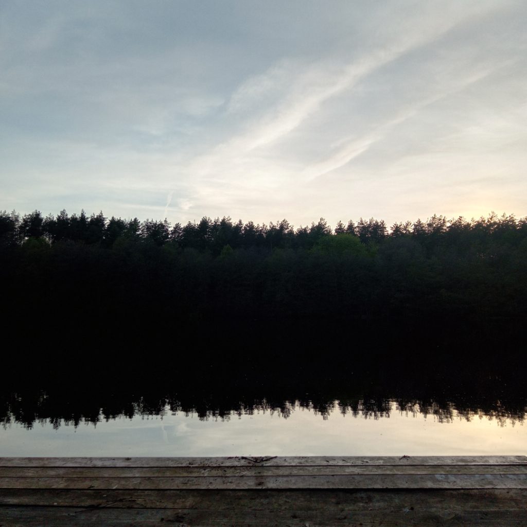 Kolorowa fotografia. Widok na taflę wody, w tle wysokie drzewa, na pierwszym planie drewniany pomost.