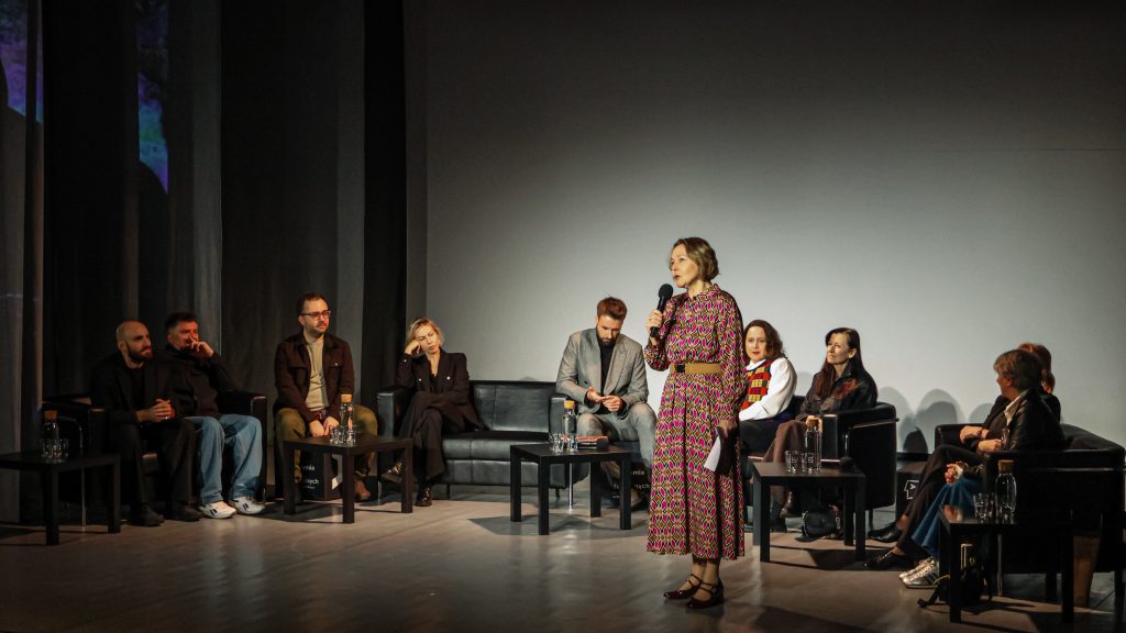 Pani dr hab. Beata Guczalska, prowadząca spotkanie stoi na scenie w sali teatralnej i jest skierowana w stronę publiki z mikrofonem w ręce. Ubrana jest w długą, wzorzystą sukienkę i buty na obcasie. Za nią siedzi reszta uczestników spotkania.
