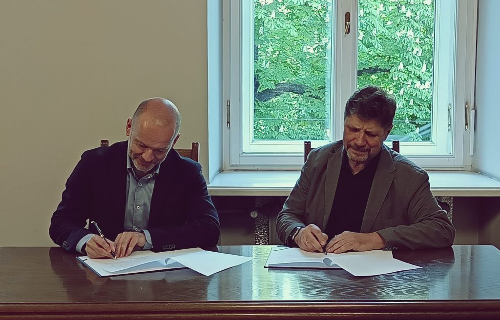 Dwóch mężczyzn – Dyrektor Talarczyk i Rektor Mielczarek – siedzi przy drewnianym stole i podpisuje dokumenty w jasnym pomieszczeniu z dużym oknem w tle, przez które widać zielone liście drzew.