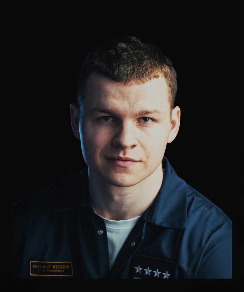 Portret. Młody ciemnowłosy mężczyzna w granatowym, rozpiętym kombinezonie i białym podkoszulku.