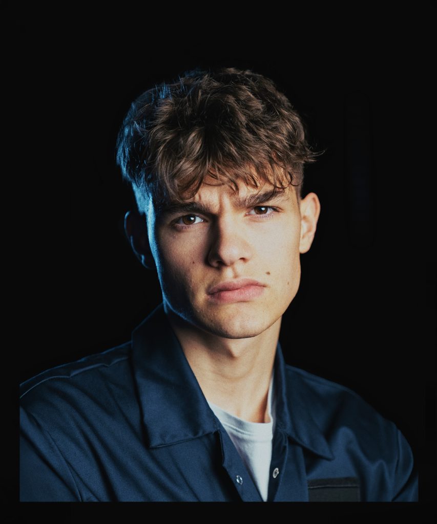 Portret. Młody, ciemnowłosy mężczyzna z brązowymi oczami. Ubrany w biały podkoszulek i granatowy kombinezon