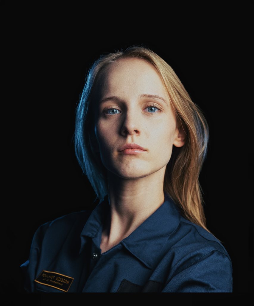 Portret długowłosej blondynki z niebieskimi oczami. Ubrana w grantowy kombinezon, na którym widnieje czarno-złota naszywka.