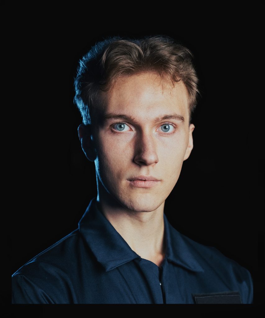 Portret młodego jasnowłosego mężczyzny o błękitnych oczach. Ubrany w granatowy kombinezon z czarną naszywką.