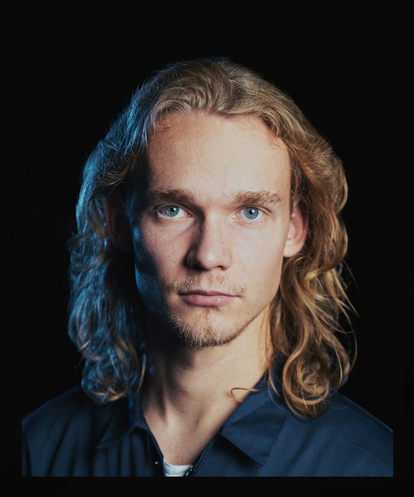 Portret. Młody blondyn z włosami długimi do ramion. Ubrany w niebieski kombinezon