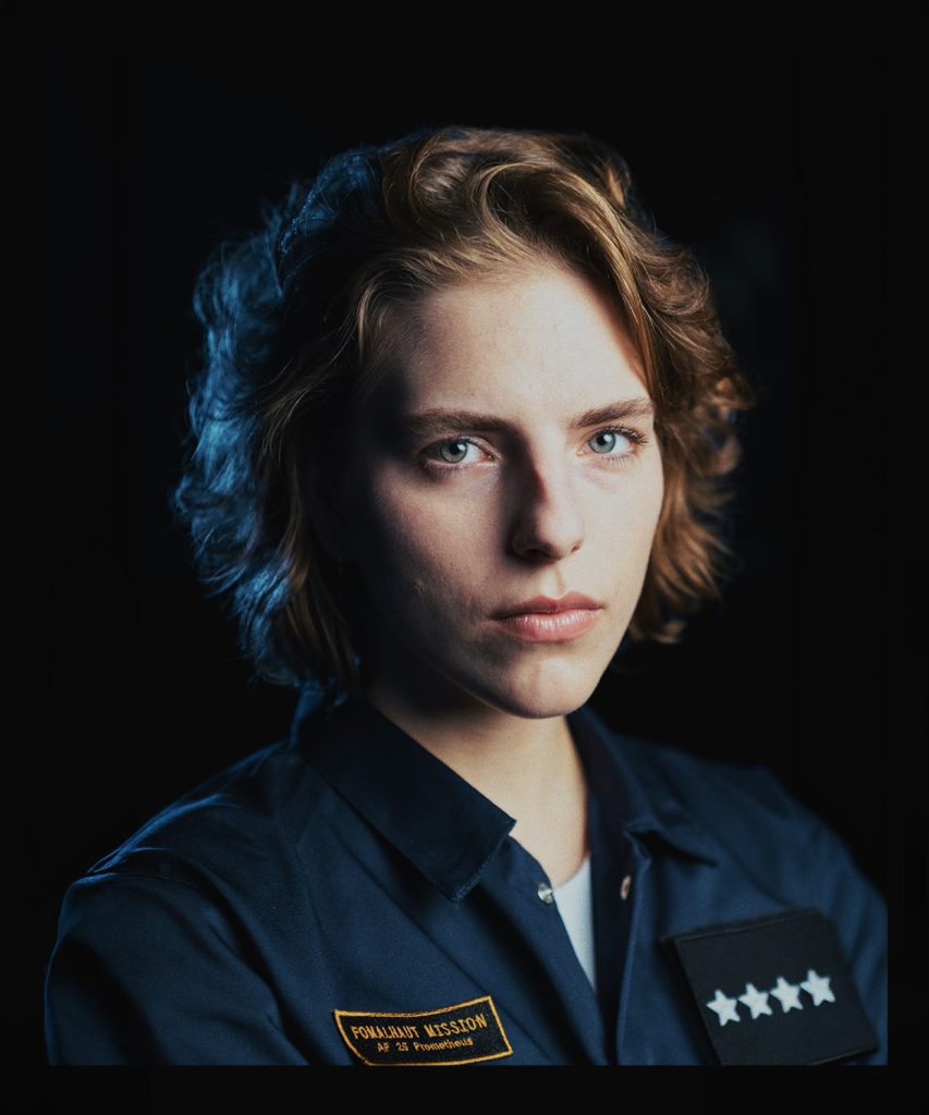 Portret młodej kobiety o jasnych, krótkich włosach. Ubrana w biały podkoszulek i granatowy kombinezon. Na kombinezonie naszywki: jedna z czterema gwiazdkami, druga czarno-złota.