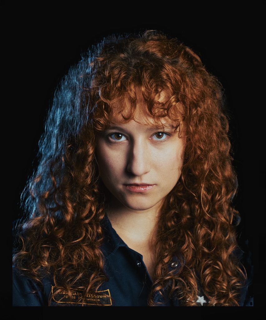 Portret młodej, rudowłosej kobiety ubranej w granatowy kombinezon. Na kombinezonie czarno-złota naszywka i naszywka z gwiazdką.