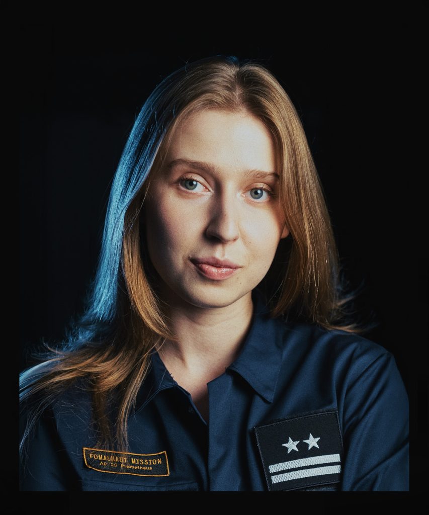 Portret młodej blondynki o szaroniebieskich oczach. Ubrana w grantowy kombinezon z dwiema naszywkami, na jednej dwie gwiazdki i dwie poziome belki, druga naszywka czarno-złota