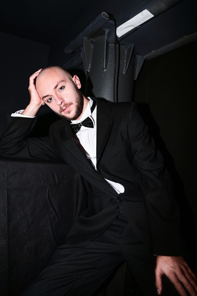 Młody mężczyzna z ciemną brodą i wąsami. Ubrany w białą koszulę i czarny garnitur, na szyi zawiązana czarna muszka. Siedzi w niewielkim, czarnym pomieszczeniu, opiera się o czarną kolumnę, ręką podpiera głowę.