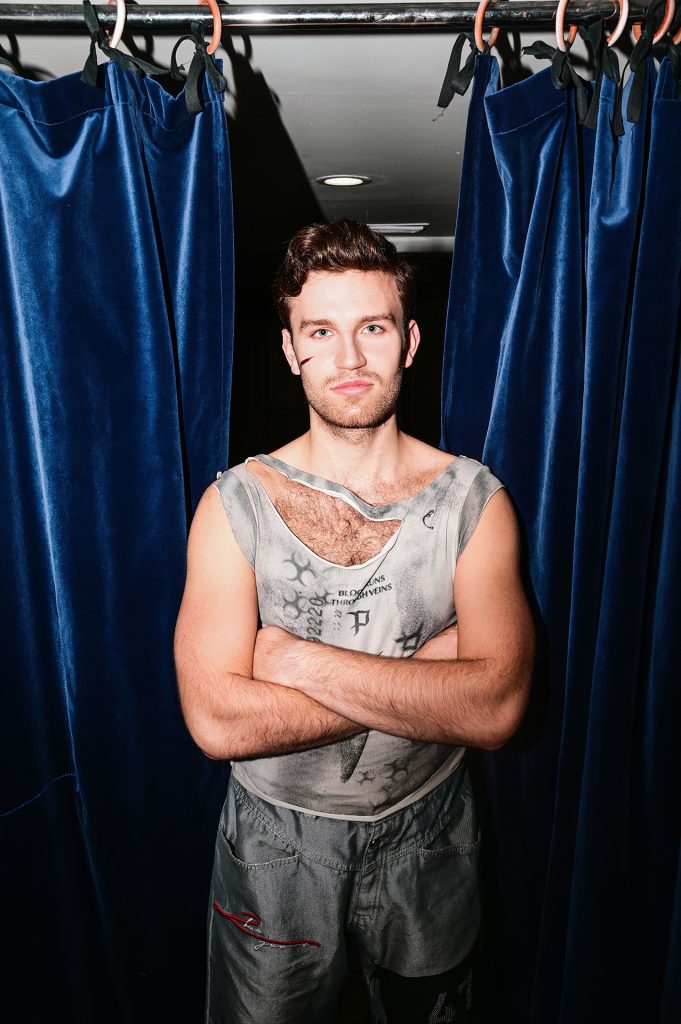 Młody, ciemnowłosy, muskularny mężczyzna w szarym, porwanym na torsie podkoszulku bez rękawów. Stoi z rękami skrzyżowanymi na piersiach. Na szarych spodniach na udzie ma naszyty numer 41 na czarnym tle. Za jego plecami ciemnogranatowa kotara. W tle biały sufit.