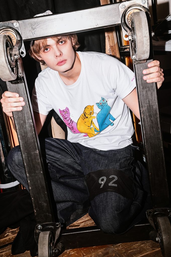 Młody chłopak w białym podkoszulku z grafiką przedstawiającą trzy koty (różowego, żółtego i niebieskiego) i ciemnych dżinsach. Na spodniach, na udzie ma opaskę z numerem 92. Chłopak klęczy, kolanem i rękami podtrzymuje ciemną ramę jakby do obrazu, do której przymocowane są kółka.