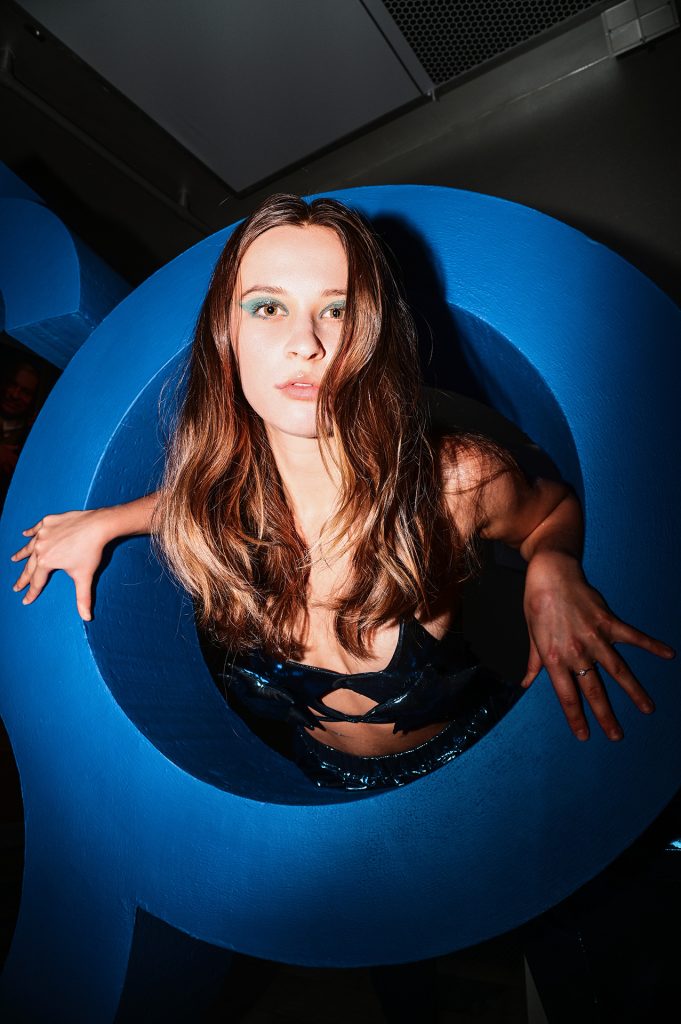 Młoda kobieta z długimi, ciemnymi, rozpuszczonymi włosami. Ubrana w ciemnoniebieski stanik w kształcie dwóch gwiazd i połyskliwe niebieskie spodenki. Jej tułów znajduje się wewnątrz dużego, niebieskiego, okrągłego walca przypominającego literę "O" i cyfrę zero.