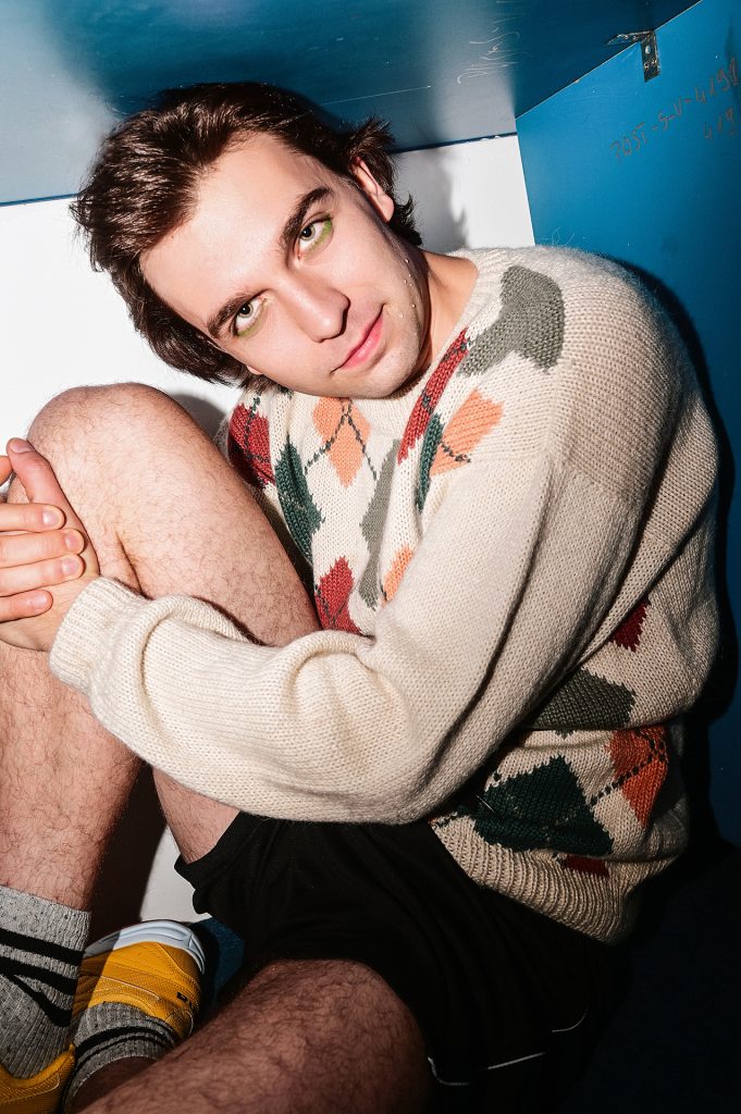 Młody ciemnowłosy mężczyzna siedzi skulony pod niebieskim biurkiem lub szafką. Pod oczami ma zielony makijaż. Ubrany w jasny sweter z wielokolorowymi geometrycznymi wzorami, czarne getry, szare skarpetki i żółte, sportowe buty. Trzyma się obiema rękami za kolano.