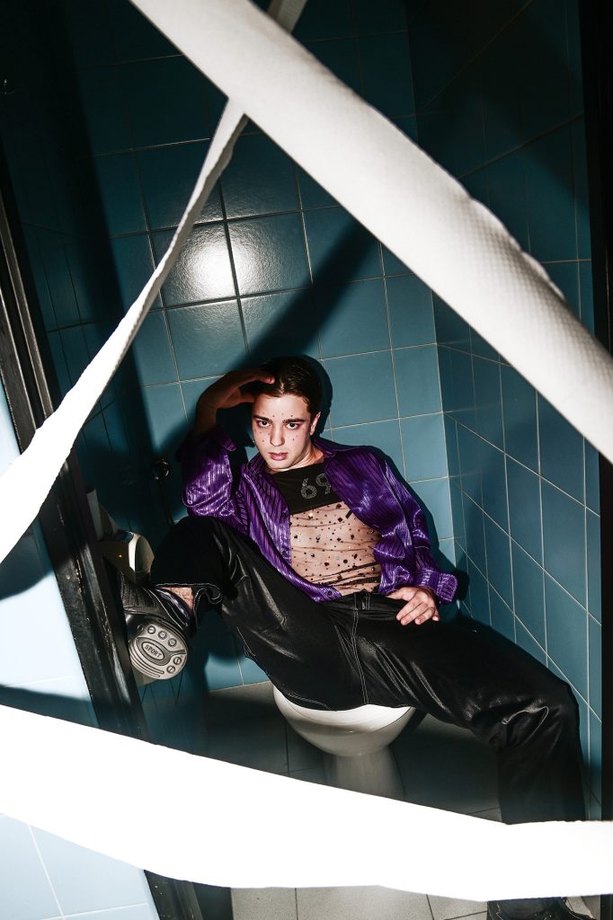 Młody, ciemnowłosy mężczyzna w fioletowej koszuli, przezroczystym podkoszulku, ciemnych spodniach i butach. Pod szyją ma czarną naszywkę z numerem 69. Siedzi na muszli klozetowej z szeroko rozstawionymi nogami, jedną nogę opiera o futrynę drzwi. Jedną ręką trzyma na udzie, drugą na czole. Wokół niego wielka wstęga papieru toaletowego ułożona w ogromny trójkąt sięgający wysoko powyżej jego głowy.