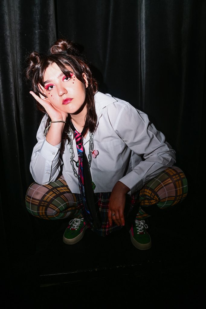Młoda ciemnowłosa kobieta w białej koszuli, spodniach w wielokolorową kratę i zielonych butach. Na powiekach ma czerwono różowy makijaż, z boku, pod oczami trzy czerwone kropki. Na szyi ma zawieszony czarno-różowo-niebieski krawat i łańcuch.