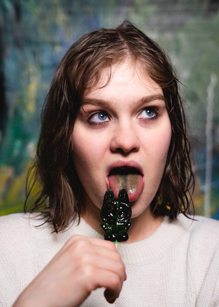 Portret. Młoda kobieta z krótkimi ciemnymi włosami, w białym podkoszulku. W ręce trzyma ciemnozielonego lizaka, na jej języku ciemnozielone ślady.