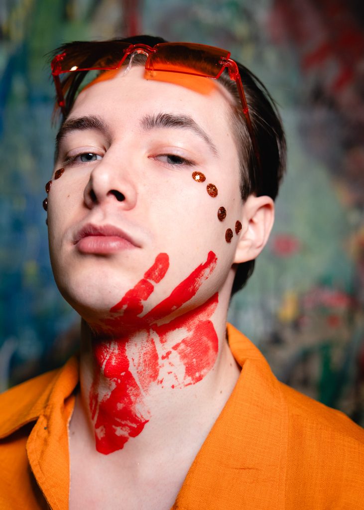 Portret. Młody, krótkowłosy brunet. Ubrany w pomarańczową marynarkę. Ma oparte o czoło pomarańczowe okulary. Na policzkach, pod oczami ma przyklejone brązowo-pomarańczowe kryształki. Na brodzie i szyi chłopaka widać krwistoczerwony odcisk dłoni.
