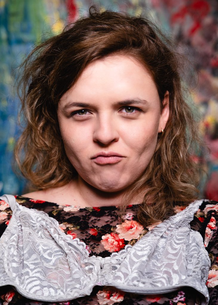Portret. Młoda kobieta z krótkimi ciemnymi włosami. Ubrana w ciemną bluzkę z motywem róż. Na jej klatce piersiowej leży zawieszony na ramionach biały biustonosz