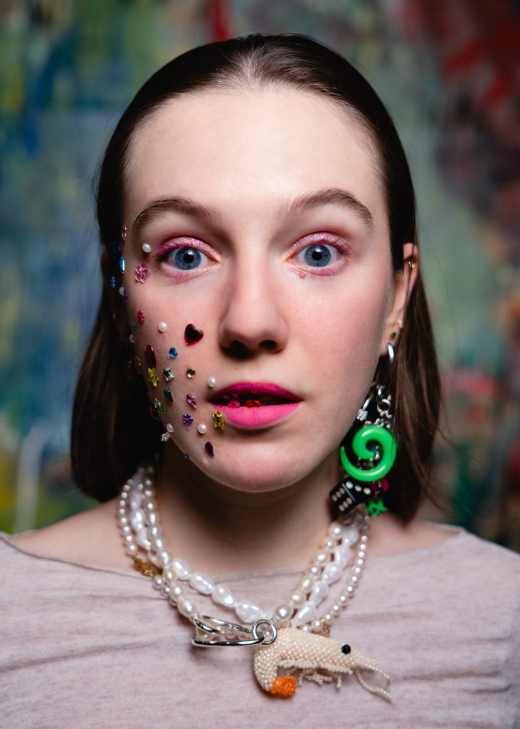 Portret. Młoda, ciemnowłosa kobieta w jasnej bluzce. Na prawym policzku i koło prawego oka ma przyklejone kryształki o różnych wzorach: serduszka, perełki, łezki, kwiatuszki. Na wargach przyklejone różowe kryształki, w uszach kolczyk: kostka do gry, zielona spirala. Kobieta ma na szyi kilka sznurów pereł